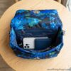 Vista aérea del interior abierto del bolso Desigual Camoflower Zurich azul floral, mostrando su capacidad para un smartphone, tarjetero, llaves y labial en sus compartimentos con cremallera.