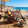 Bolso grande Desigual diseñado por Christian Lacroix con estampado floral sobre fondo negro, apoyado en una mesa de madera de una cafetería frente al mar. Escena veraniega con café y planta suculenta que resalta el diseño artístico. Disponible en amathingshop.com.