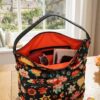 Primer plano del bolso hobo Desigual Christian Lacroix floral negro, medio abierto sobre una mesa de madera. El interior naranja brillante está lleno con una cartera, gafas de sol, libreta, pintalabios y móvil, mostrando su gran capacidad y organización. Luz natural de una ventana. Disponible en amathingshop.com.