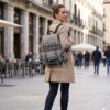 Mujer con abrigo beige caminando por una ciudad europea con la mochila Desigual Janis Tribeca gris jaspeada, detalle bordado LOVE y correas ajustables, disponible en amathingshop.com