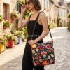 Mujer elegante caminando por una calle adoquinada de un pueblo francés con un vestido negro, llevando al hombro el bolso grande floral Desigual Christian Lacroix Pekin. La imagen muestra el tamaño real y la versatilidad del bolso. Disponible en amathingshop.com.