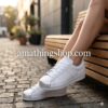 Primer plano de zapatillas Adidas Superstar blancas con puntera de concha plateada metalizada, puestas por una chica sentada en un banco de madera, imagen de amathingshop.com.