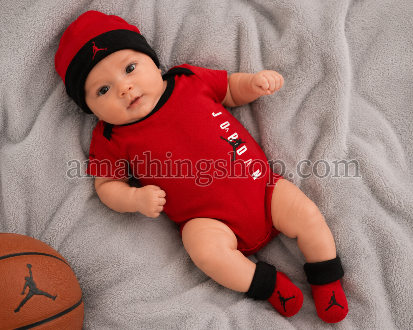 Bebé recién nacido con conjunto Jordan rojo compuesto por body, gorro y patucos sobre manta gris suave.