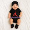 Bebé sonriendo con conjunto oficial Nike Jordan para bebé compuesto por body negro, gorro y patucos con logotipo Jumpman rojo modelo MJ0041-023