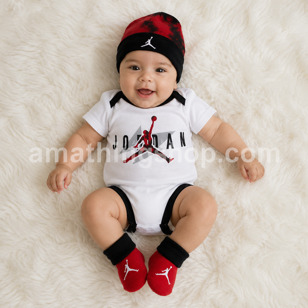 Bebé sonriendo con conjunto original Nike Air Jordan de tres piezas formado por body blanco, gorro rojo y negro y patucos a juego modelo NJ0446-001