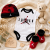 Conjunto Nike Air Jordan para bebé con body blanco, gorro rojo y negro y patucos a juego sobre una manta de punto junto a un peluche y accesorios de baby shower modelo NJ0446-001
