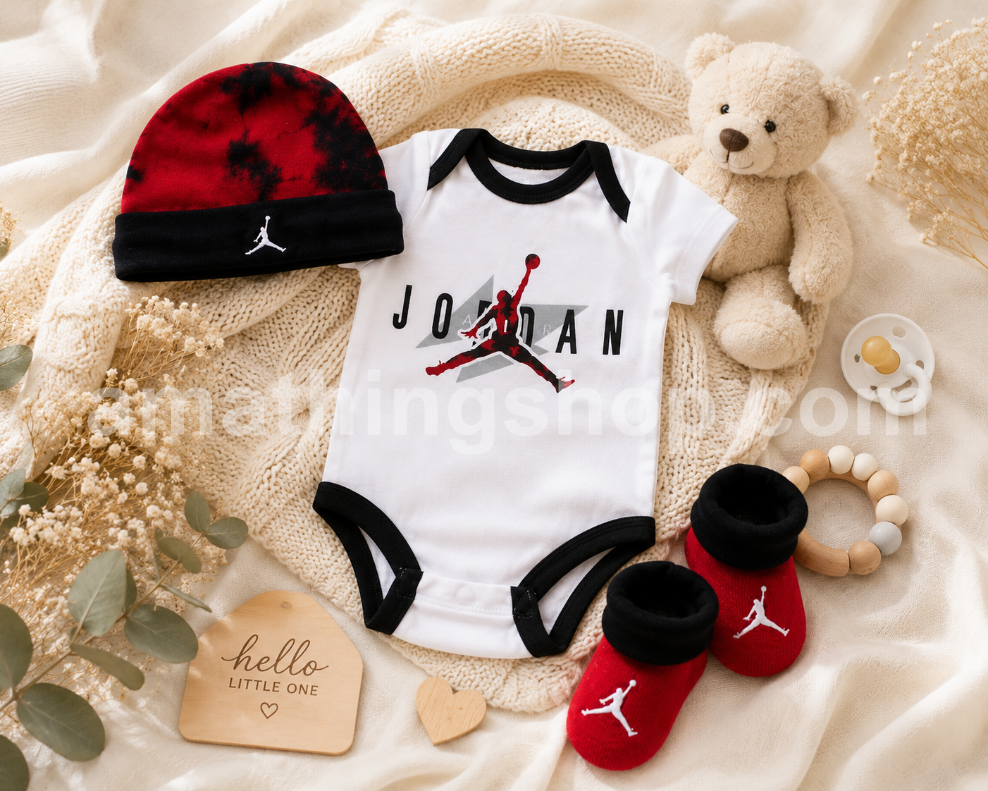 Conjunto Nike Air Jordan para bebé con body blanco, gorro rojo y negro y patucos a juego sobre una manta de punto junto a un peluche y accesorios de baby shower modelo NJ0446-001
