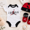 Body Nike Air Jordan para bebé con gorro rojo y negro y patucos a juego sobre fondo de manta blanca con peluche y chupete modelo NJ0446-001