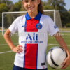 Niña con camiseta oficial Nike Paris Saint-Germain 2020/21 blanca posando en campo de fútbol con balón. Equipación visitante PSG infantil con diseño rojo y azul ideal para niñas aficionadas al fútbol.