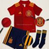Vista detallada del conjunto de fútbol infantil de España, mostrando la calidad del escudo, el tejido de la camiseta roja y el pantalón azul marino en estilo catálogo profesional