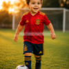Niño pequeño corriendo con equipación roja de España y balón de fútbol en campo al atardecer estilo lifestyle premium