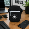 Bandolera Converse negra sobre escritorio lifestyle urbano Bandolera Converse negra tipo crossbody sobre mesa moderna con portátil, gafas e iPhone.