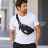Hombre con camiseta blanca llevando riñonera Jordan Jumpman negra 9A0623-023 cruzada al pecho, con logotipo blanco y correa ajustable. Copiar ALT