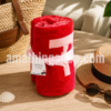 Toalla Naf Naf roja enrollada sobre mesa de madera con bolso de playa, sombrero y gafas de sol. Toalla de algodón ideal para verano, piscina y vacaciones.