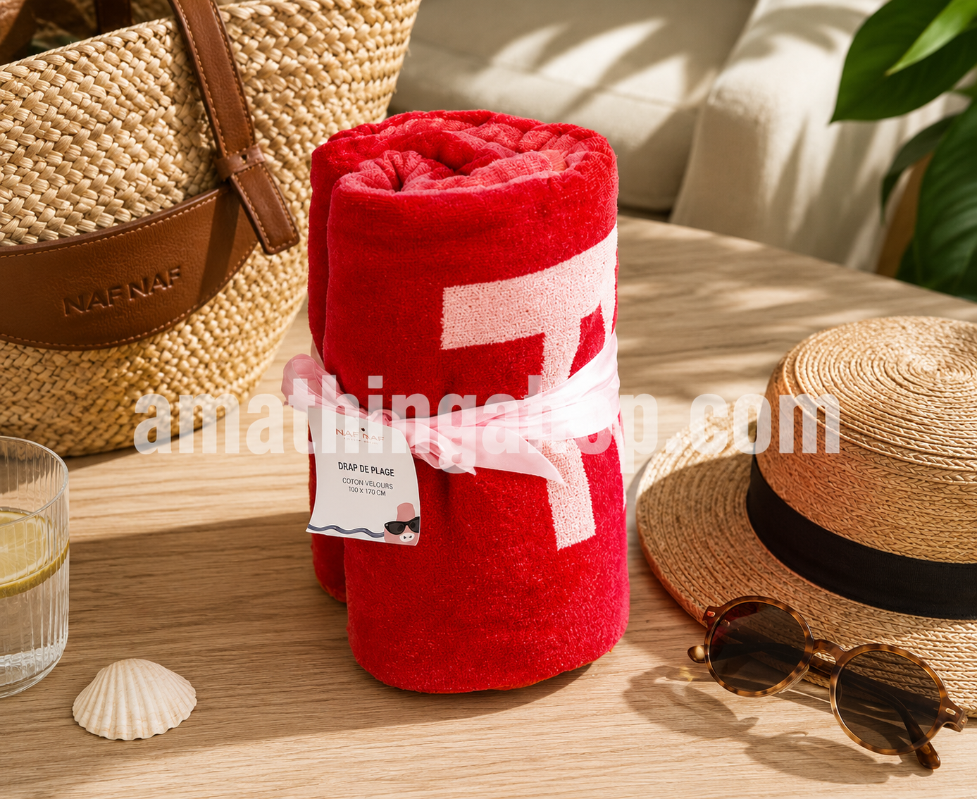 Toalla Naf Naf roja enrollada sobre mesa de madera con bolso de playa, sombrero y gafas de sol. Toalla de algodón ideal para verano, piscina y vacaciones.