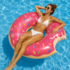 Mujer relajándose sobre flotador donut fresa rosa de 110 cm aproximadamente en una piscina azul durante un día de verano, con diseño divertido tipo donut mordido y web Amathingshop.com integrada.