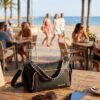 Bolso Desigual Prime Urus Maxi en Terraza frente al Mar Bolso bandolera mediano Desigual Prime Urus Maxi negro con bordados rosas y naranjas, apoyado en una mesa de madera en una terraza soleada frente a la playa. Se aprecian unas gafas de sol y un móvil junto al bolso, con palmeras y gente paseando de fondo. Estilo casual chic disponible en amathingshop.com.