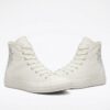 zapatillas-converse-blancas-bordado-floral Primer plano de un par de zapatillas Converse de lona blancas de caña alta con un bordado floral gris plateado en el lateral, sobre un fondo blanco.