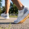 Primer plano de las zapatillas Reebok Lite Plus 2.0 para hombre en color blanco y gris mientras camina por asfalto. Se detalla la media suela de EVA para amortiguación superior y la suela de goma resistente. Calzado deportivo ligero talla 44 disponible en amathingshop.com.