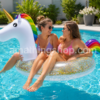 Dos jóvenes disfrutando de un flotador unicornio blanco con purpurina dorada, cuerno dorado y melena arcoíris en una piscina durante un día soleado de verano.