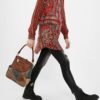 Mochila bolso Desigual Atica Positano llevada en la mano por una mujer con look urbano, camisa estampada roja, vaqueros negros y botas negras. Imagen lifestyle que muestra el tamaño real aproximado, el diseño patchwork marrón y denim, la asa superior y el estilo casual de esta mochila convertible Desigual para mujer.