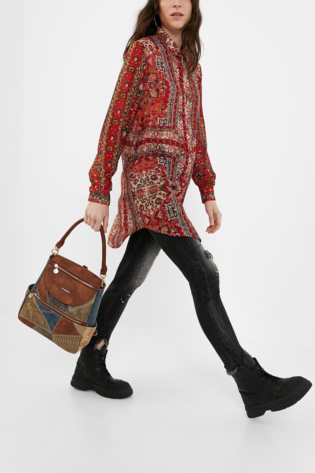 Mochila bolso Desigual Atica Positano llevada en la mano por una mujer con look urbano, camisa estampada roja, vaqueros negros y botas negras. Imagen lifestyle que muestra el tamaño real aproximado, el diseño patchwork marrón y denim, la asa superior y el estilo casual de esta mochila convertible Desigual para mujer.
