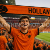 Aficionado Levantando la Bufanda Oficial Adidas Países Bajos Holanda FJ0994 en el Estadio Aficionado levantando la bufanda oficial Adidas Países Bajos Holanda FJ0994 naranja y negra con diseño HOLLAND durante un partido de fútbol en un estadio