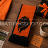 Bufanda Oficial Adidas Países Bajos Holanda FJ0994 como Regalo para Aficionados al Fútbol Bufanda oficial Adidas Países Bajos Holanda FJ0994 naranja y negra sobre una mesa junto a una camiseta, regalo y accesorios de aficionado al fútbol
