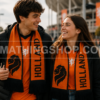 Pareja de Aficionados con Bufanda Oficial Adidas Países Bajos Holanda FJ0994 Hombre y mujer jóvenes llevando la bufanda oficial Adidas Países Bajos Holanda FJ0994 naranja y negra con diseño reversible durante un partido de fútbol
