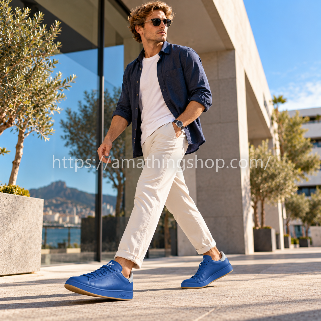 Hombre caminando con zapatillas Adidas Stan Smith Recon azules H06186, pantalones beige y camisa azul marino en una plaza urbana.