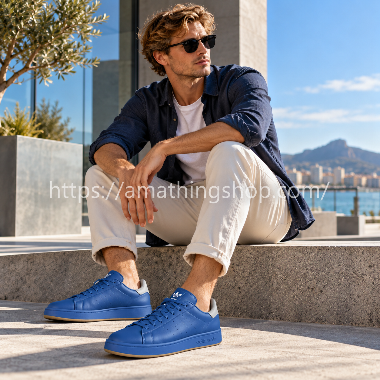 Hombre rubio con zapatillas Adidas Stan Smith Recon azules H06186, pantalones beige y camisa azul en un entorno urbano junto al mar.