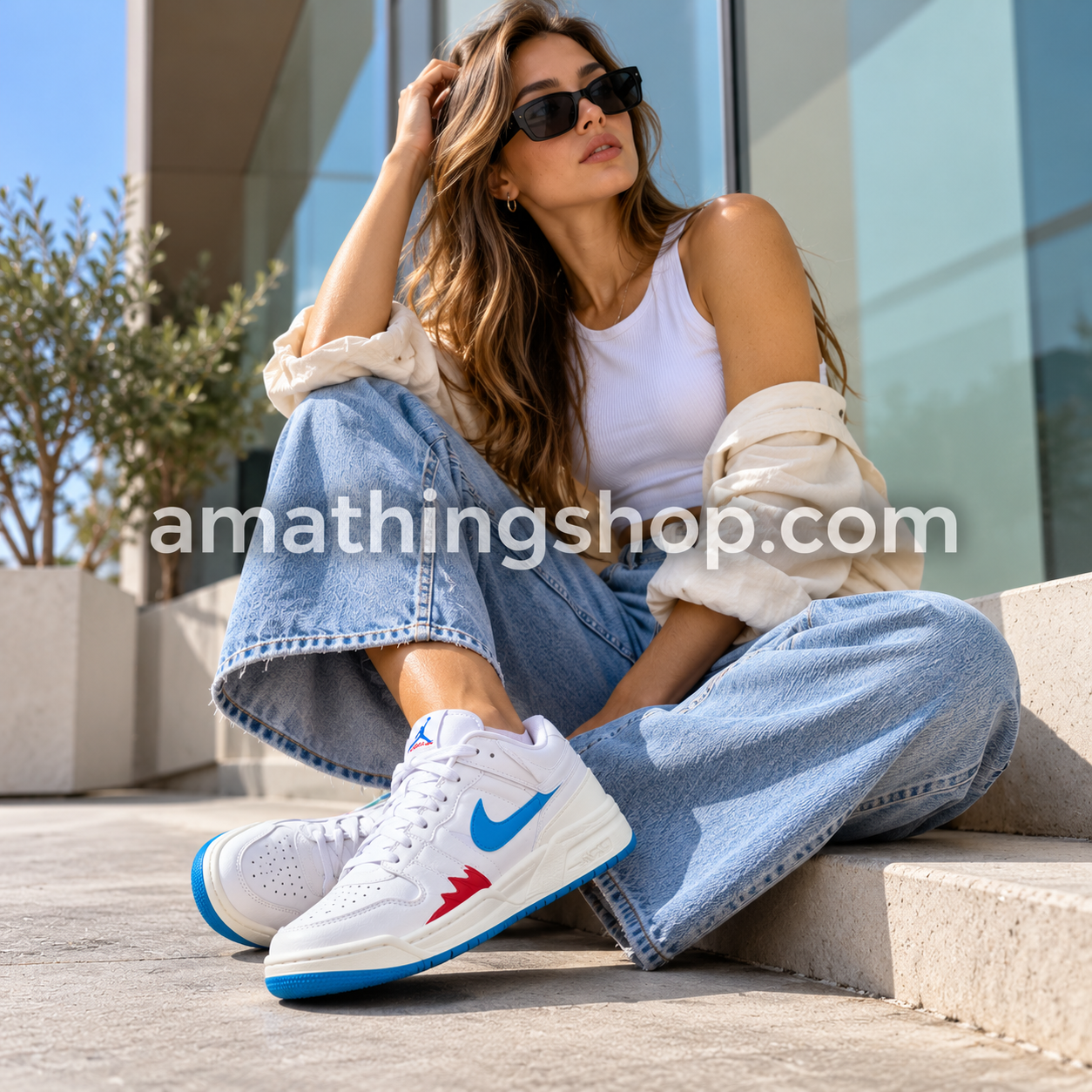 Mujer con gafas de sol, vaqueros anchos y zapatillas Jordan Stadium 90 blancas con Swoosh azul y detalles rojos, sentada en una terraza urbana.